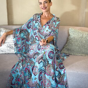 May include: A flowing, light blue paisley print dress with a V-neck and wide sleeves. The dress features a vibrant pattern of pink, purple, and teal paisley designs. A tassel hangs from the neckline. The dress is worn while seated on a gray sofa.