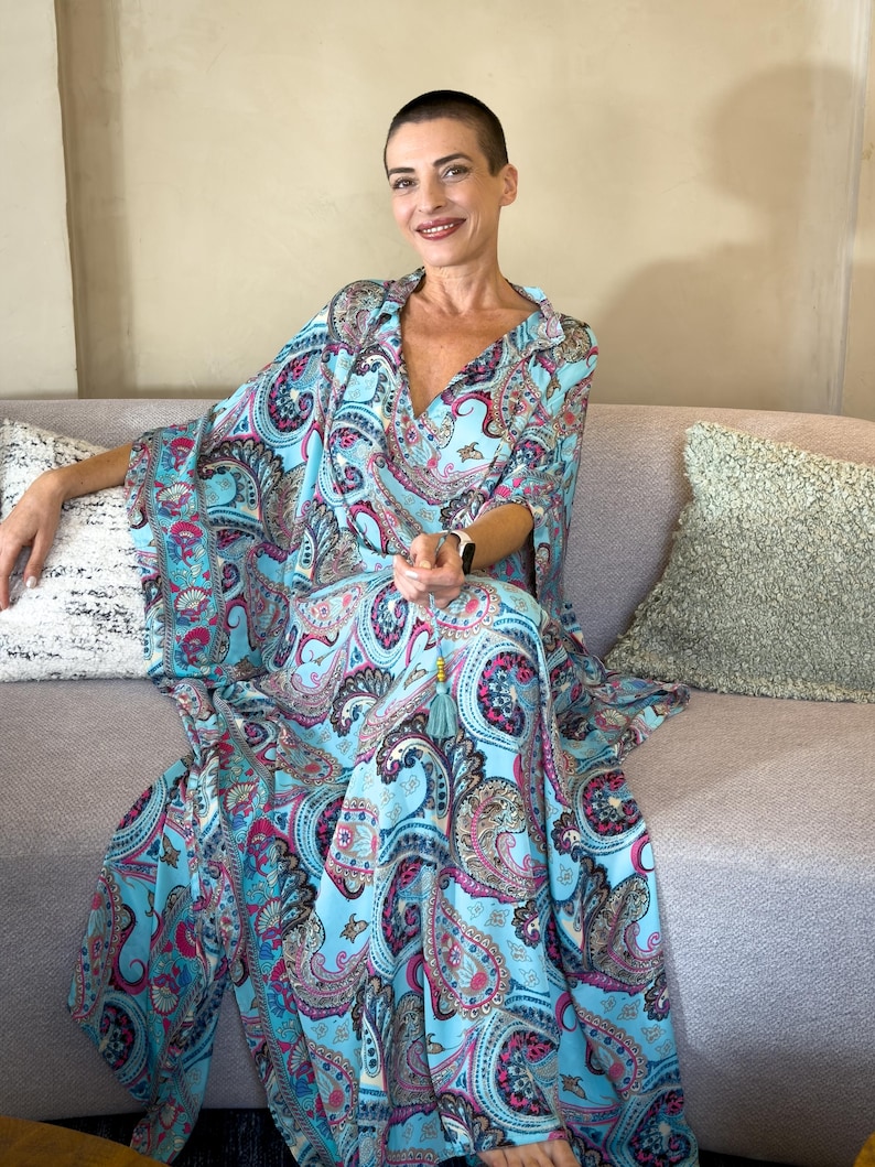 May include: A flowing, light blue paisley print dress with a V-neck and wide sleeves. The dress features a vibrant pattern of pink, purple, and teal paisley designs. A tassel hangs from the neckline. The dress is worn while seated on a gray sofa.