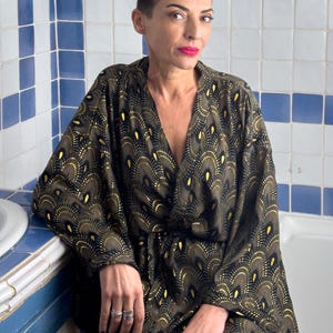 May include: A woman wearing a black and gold kimono with a peacock feather design. The kimono has a V-neck and long sleeves. The woman is sitting in a bathroom with white and blue tiles.
