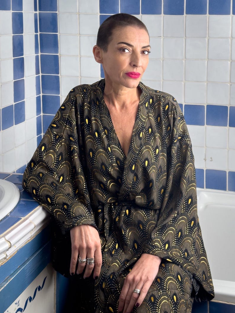 May include: A woman wearing a black and gold kimono with a peacock feather design. The kimono has a V-neck and long sleeves. The woman is sitting in a bathroom with white and blue tiles.