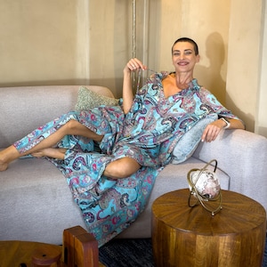 May include: A light blue paisley print robe and matching pants. The robe has a V-neck and long sleeves. The person is sitting on a gray sofa with a decorative pillow. A gold-colored globe decoration sits on a wooden table.
