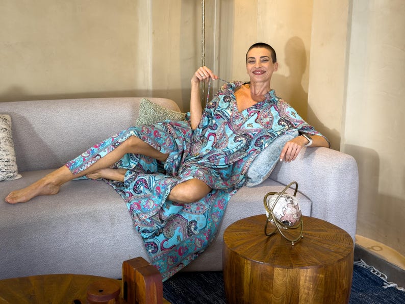 May include: A light blue paisley print robe and matching pants. The robe has a V-neck and long sleeves. The person is sitting on a gray sofa with a decorative pillow. A gold-colored globe decoration sits on a wooden table.