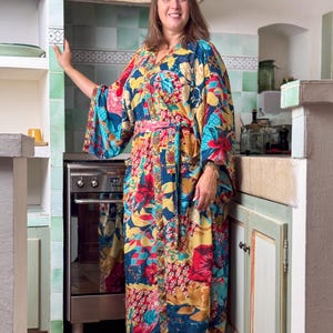 May include: A woman wearing a long, colourful floral print kimono with a pink sash. The kimono has a blue background with red, yellow, and green flowers. She is standing in a kitchen with a stove and cabinets in the background.