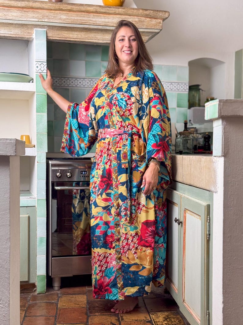May include: A woman wearing a long, colourful floral print kimono with a pink sash. The kimono has a blue background with red, yellow, and green flowers. She is standing in a kitchen with a stove and cabinets in the background.