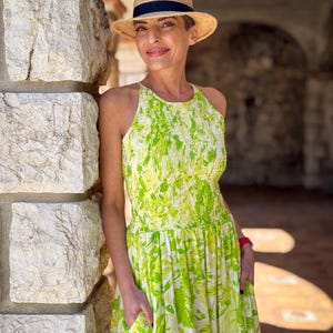 Può includere: Una donna indossa un cappello di paglia con una fascia blu navy e un vestito all'americana. L'abito presenta un motivo floreale verde e bianco. La donna è appoggiata a un muro di pietra, con le mani in tasca.