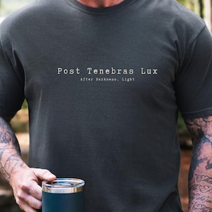 May include: Dark gray t-shirt with the text "Post Tenebras Lux" and "After Darkness, Light" printed on the front. A person is holding a dark blue mug with a silver rim. The image is set outdoors with trees in the background.
