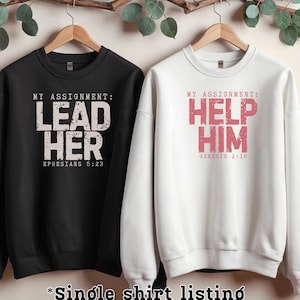 May include: Two long-sleeved sweatshirts, one black and one white, hanging on wooden hangers. The black sweatshirt has the text "MY ASSIGNMENT: LEAD HER EPHESIANS 5:23" in white. The white sweatshirt has the text "MY ASSIGNMENT: HELP HIM GENESIS 2:18" in red.