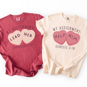 May include: Two t-shirts, one burgundy and one cream, are displayed. The burgundy shirt has the text "My Assignment: Lead Her" with "Ephesians 5:23" below. The cream shirt reads "My Assignment: Help Him" with "Genesis 2:18".