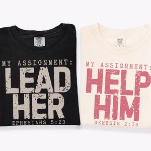 May include: Two folded t-shirts, one black and one cream. The black shirt has the text "MY ASSIGNMENT: LEAD HER" and "EPHESIANS 5:23". The cream shirt says "MY ASSIGNMENT: HELP HIM" and "GENESIS 2:18".