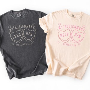 May include: Two t-shirts with text and heart graphics. One is charcoal gray with the words "MY ASSIGNMENT: LEAD HER" and "EPHESIANS 5:23". The other is cream-colored with "MY ASSIGNMENT: HELP HIM" and "GENESIS 2:18". Both shirts have a vintage, worn look.