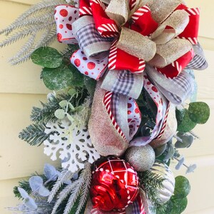 Puede incluir: Una guirnalda navide&ntilde;a festiva con un gran lazo rojo, blanco y plateado, adornada con adornos rojos y plateados y copos de nieve blancos. La guirnalda est&aacute; decorada con vegetaci&oacute;n y ramas de pino escarchadas.