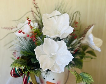 May include: A white snowman-shaped planter with a red and green plaid bow, filled with white flowers and greenery.