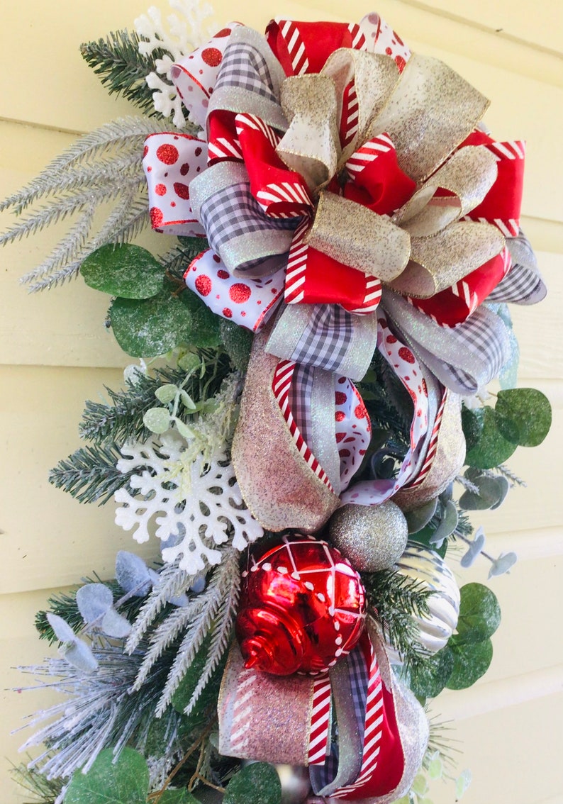 Puede incluir: Una guirnalda navide&ntilde;a festiva con un gran lazo rojo, blanco y plateado, adornada con adornos rojos y plateados y copos de nieve blancos. La guirnalda est&aacute; decorada con vegetaci&oacute;n y ramas de pino escarchadas.