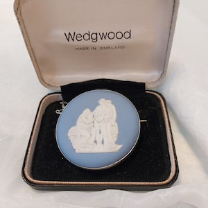 May include: A Wedgwood cameo brooch in a blue and white color scheme, featuring a white cameo of two figures on a blue background. The brooch is in a black velvet box with a white label that reads "Wedgwood Made in England".