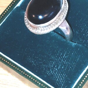 May include: A silver ring featuring a large, smooth, oval-shaped black stone set in a bezel. The metal band displays a textured, braided pattern around the stone setting, providing a detailed border for the polished, dark, reflective center piece.