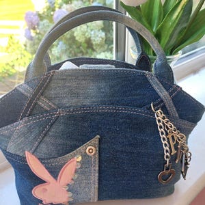 May include: A dark blue denim handbag featuring a patchwork design with visible stitching. The bag includes two top handles, a front pocket adorned with a light pink embroidered rabbit logo, and a silver-toned metal chain charm with a letter P and heart.