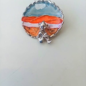 May include: A silver pin with a small, silver figurine of a person sitting on a round, painted enamel disc. The disc has a blue, white, and orange design.