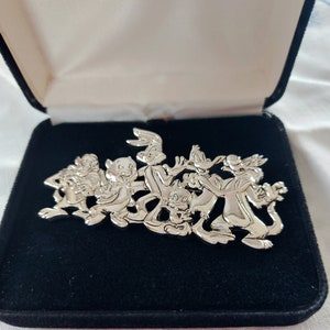 May include: A silver-toned brooch featuring a group of cartoon characters, including Bugs Bunny, Daffy Duck, Tweety Bird, Sylvester the Cat, and Porky Pig. The characters are depicted in a playful, animated style, with Bugs Bunny holding a carrot and Daffy Duck holding a gun.