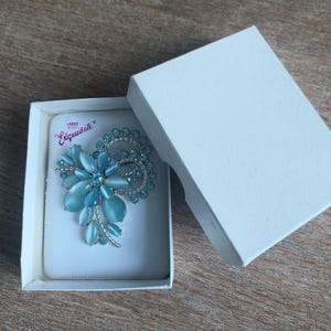 May include: A blue and silver brooch in the shape of a flower with the text "Exquisite" on the white card it is displayed on.