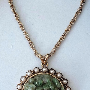 May include: Gold-tone necklace featuring a round pendant with a green stone center. The pendant is framed by small faux pearls and a gold-tone border. The chain is a twisted rope design.