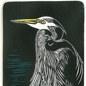 May include: A black and white woodcut print of a heron standing in tall grass. The heron has a yellow beak and a yellow eye. The background is black with a blue and green water pattern.
