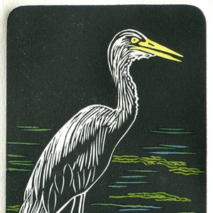 May include: A black and white woodcut print of a young heron standing on a rock in a pond. The heron is white with a yellow beak and black eyes. The pond is depicted with green and blue lines. The print is signed "Sim '22" and numbered "3/30".