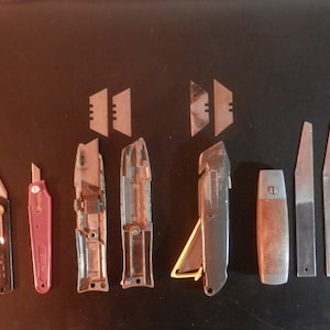 May include: A collection of utility knives and blades, including box cutters and scrapers, arranged on a dark surface. The knives vary in size, color, and design, with some featuring metal and plastic handles. Several spare blades are also present.