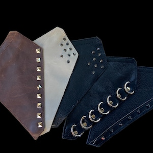 May include: Four leather wrist cuffs in brown, off-white, and black. The brown cuff has gold studs, the off-white cuff has black studs, and the black cuffs have silver grommets and studs. The cuffs are arranged on a black background.