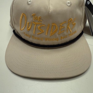 May include: A tan baseball cap with the words "THE OUTSIDERS" embroidered in gold thread. The text "Tony Award Winning Best Musical" is embroidered below the main text. The cap has a black rope detail and a curved brim.