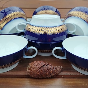 May include: Set of six blue and gold porcelain soup bowls with ornate gold trim. The bowls have two handles and are arranged on a wooden surface.