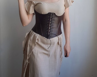 May include: A brown leather corset with a lace-up front. The corset is worn over a white, off-the-shoulder top and a long, flowing skirt.