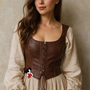 Op de afbeelding: Een bruin leren korset met een vierkante halslijn en een veter aan de voorkant. Het korset wordt gedragen over een crèmekleurige blouse met lange, pofmouwen en een bijpassende rok. De algehele stijl doet denken aan historische of renaissance mode.