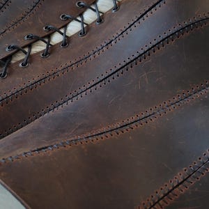 Op de afbeelding: Een donkerbruin leren korset met meerdere verticale panelen die zijn verbonden door zichtbare zwarte stiksels. Het kledingstuk heeft een reeks metalen oogjes langs de zijkant, geregen met een dun zwart koord voor aanpassing.