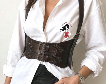 May include: A dark brown leather corset belt featuring two horizontal straps with brass-toned buckles and decorative cutouts. The belt is worn over a crisp white button-down shirt that displays a small graphic of a figure in a red corset.