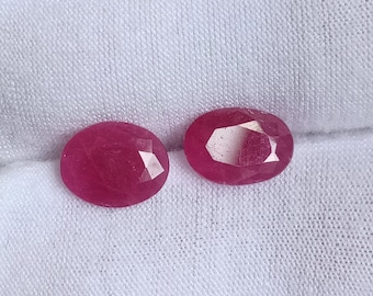 Puede incluir: Dos piedras preciosas de rubí de forma ovalada y facetadas, de un tono rosa intenso. Las piedras preciosas están colocadas sobre un fondo de tela blanca.