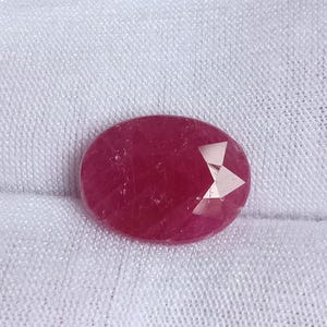 May include: A deep red oval-shaped gemstone with a faceted cut. The gemstone has a slight shimmer and is set against a white background.