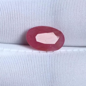 May include: Oval-shaped, faceted ruby gemstone. The ruby is a deep pinkish-red color with a lighter, geometric-shaped facet in the center. The gemstone is set against a white, textured background.