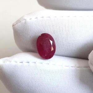May include: A faceted, oval-shaped ruby gemstone. The ruby is a deep, rich red colour and is set against a white background. The gemstone has multiple facets that reflect light, creating a sparkling effect. The ruby is a precious stone, often used in jewellery.