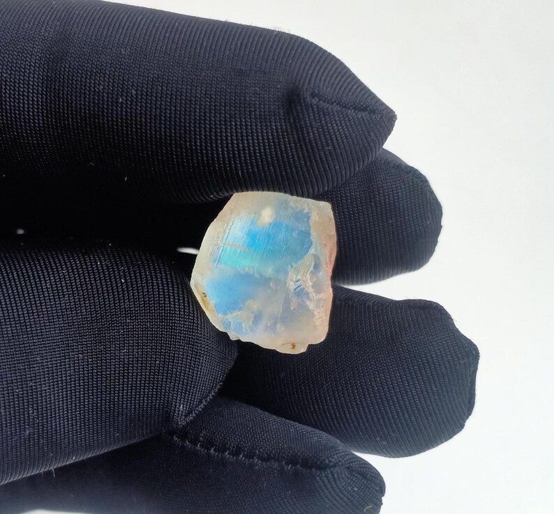 Puede incluir: Una piedra preciosa blanca, transl&uacute;cida e iridiscente con destellos azules y verdes. La piedra preciosa tiene una forma irregular y una superficie rugosa.