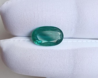 May include: An oval-shaped, emerald-green gemstone. The gem is displayed against white fabric, accentuating its clarity and colour. The gemstone's facets reflect light, producing a sparkling effect. The presentation suggests a focus on jewellery or gemstones.