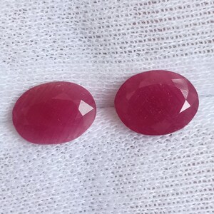 May include: Two oval-shaped, faceted gemstones with a deep pink hue. The gemstones are set against a white fabric background.