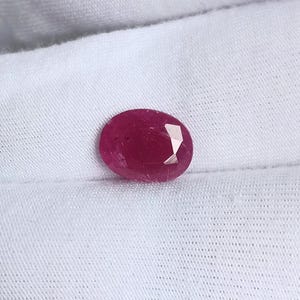 Peut inclure: Une pierre précieuse ovale de couleur rouge foncé avec des facettes, sur un fond de tissu blanc.