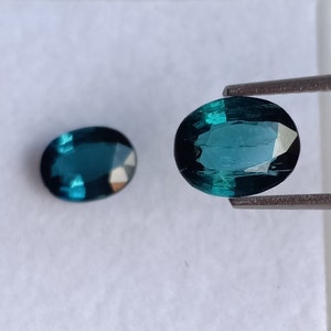 Könnte beinhalten: Zwei ovale Edelsteine, einer mit einer Pinzette gehalten. Beide sind tief blaugrün mit einem leichten Grünstich. Die Edelsteine sind facettiert und haben einen glasartigen Glanz.