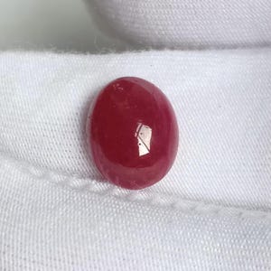 Op de afbeelding: Een dieprode ovale cabochon edelsteen, waarschijnlijk een robijn, tegen een witte stoffen achtergrond.