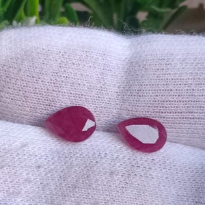 May include: Two pink pear-shaped gemstones on a white fabric background.