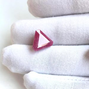 May include: A triangular gemstone in a deep pink colour. The gem is displayed against a white background, highlighting its geometric shape and polished surfaces. The facets of the gem reflect light, creating a sparkling effect.