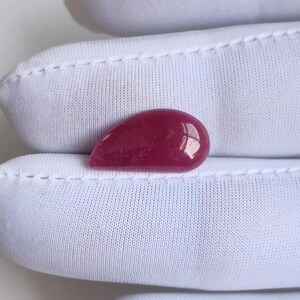 Pode incluir: Uma pedra preciosa polida em forma de gota, num tom vermelho profundo e rico. A gema tem uma superfície lisa e brilhante, que reflete a luz. A pedra está colocada sobre um fundo branco, realçando a sua cor e forma. Esta pedra preciosa é adequada para a fabricação de joias.