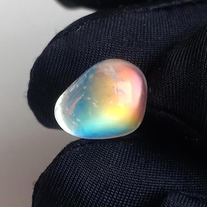 May include: A polished, iridescent gemstone with a smooth, rounded shape. The stone displays a spectrum of colors, including blue, yellow, and pink, creating a rainbow effect. The gemstone is held against a dark background.