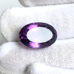 May include: An oval-cut amethyst gemstone featuring a deep purple hue with lighter violet tones and a faceted surface that reflects light. The gemstone is displayed against a white fabric background, showcasing its transparent clarity and polished finish.