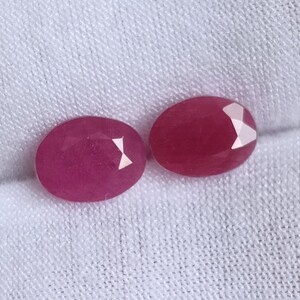 May include: Two oval-shaped, faceted ruby gemstones, both a deep pink color, are displayed on a white fabric background.